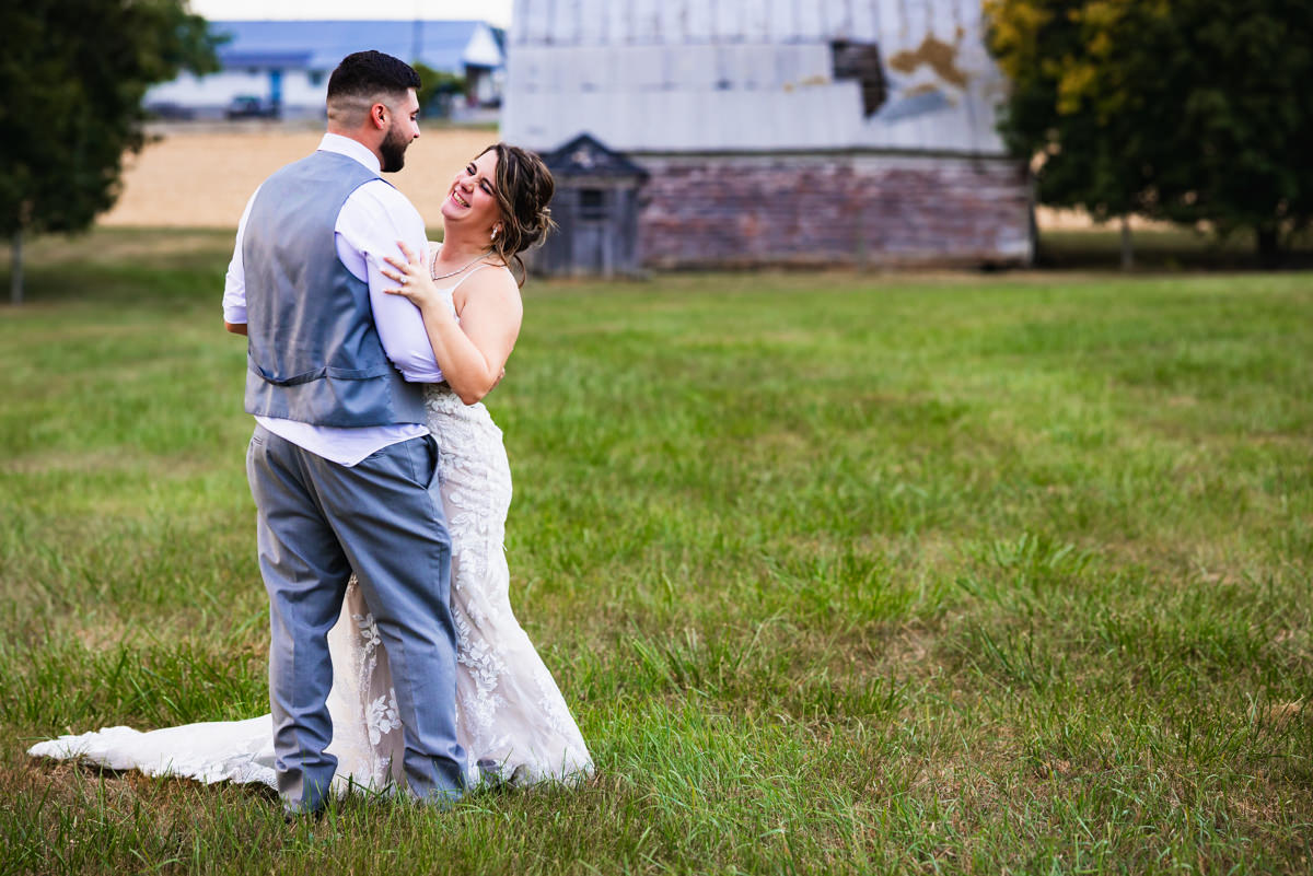 Newly married couple laughing together in open field, barn in the background.