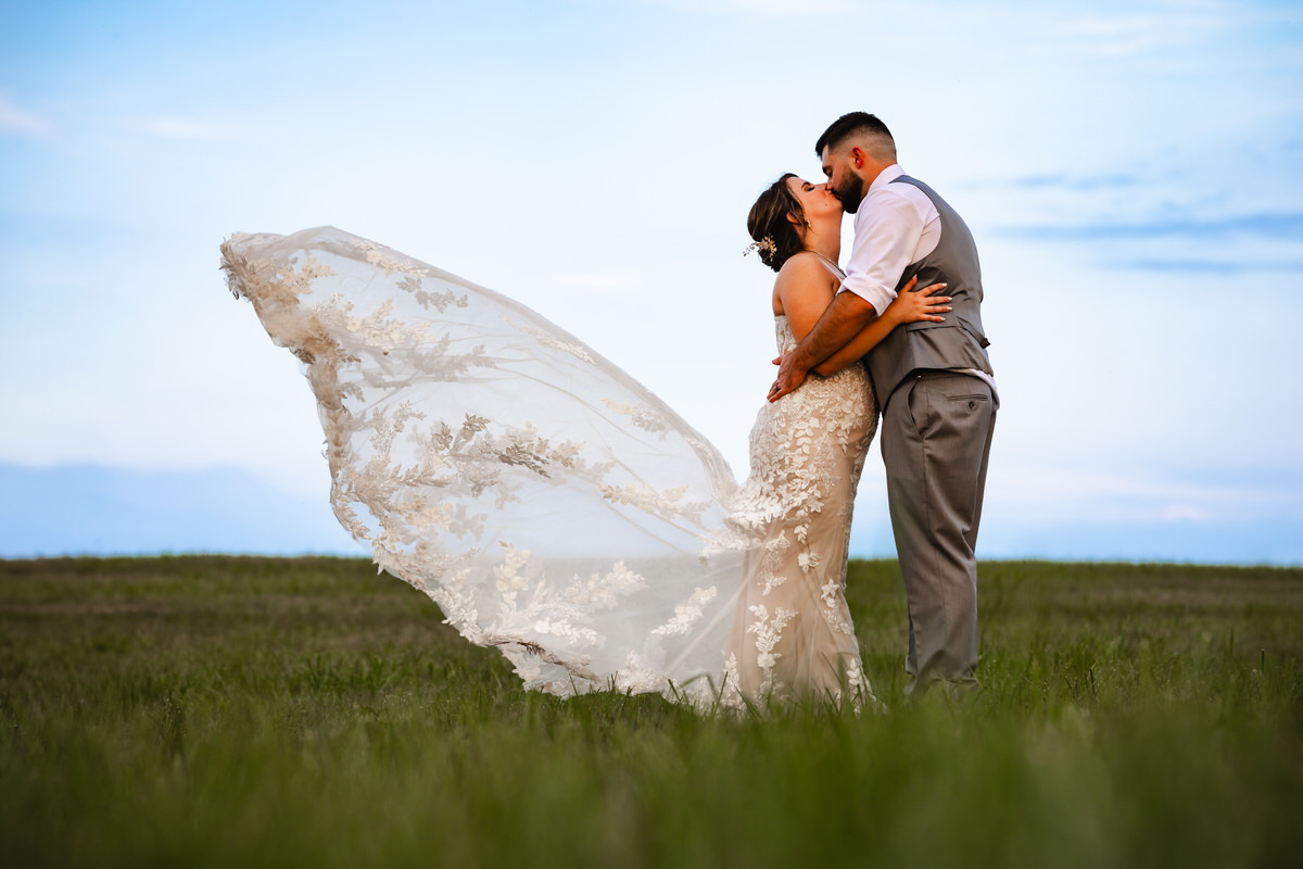 Couple kiss in open field while brides dress blows in the wind