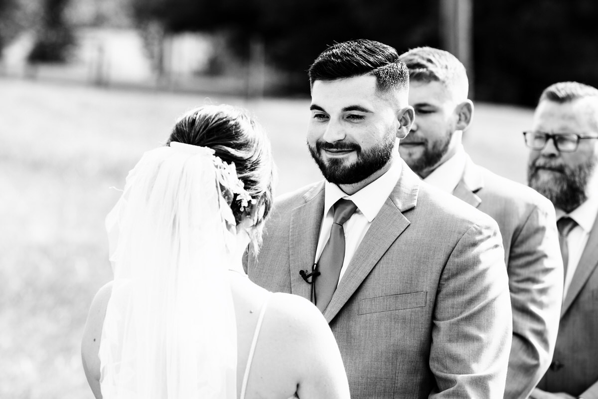 Black and white image of groom smiling at bride during their outdoor ceremony