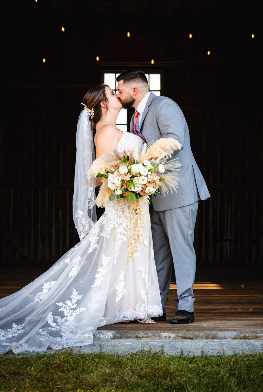 Bride and groom kiss with the barn interior blacked out behind them, window and overhanging lights set the mood
