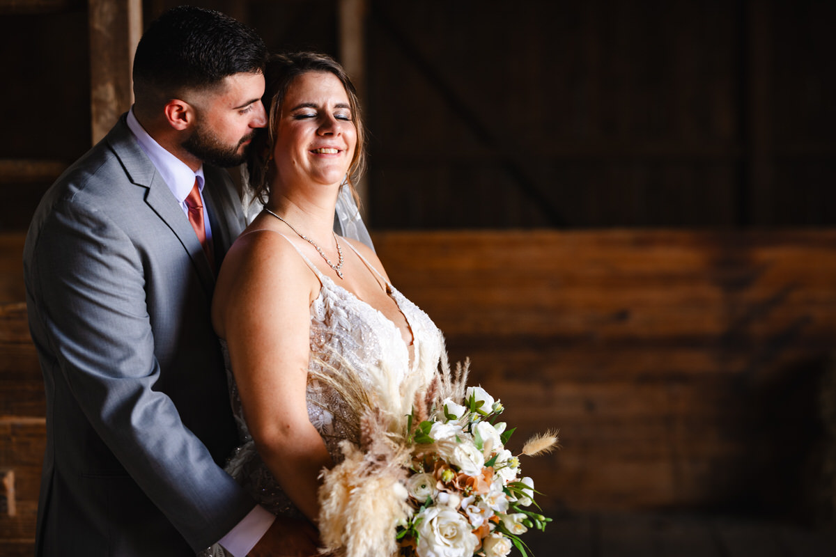 The groom is whispering into the bride's ear making her smile while they have a moment in the barn